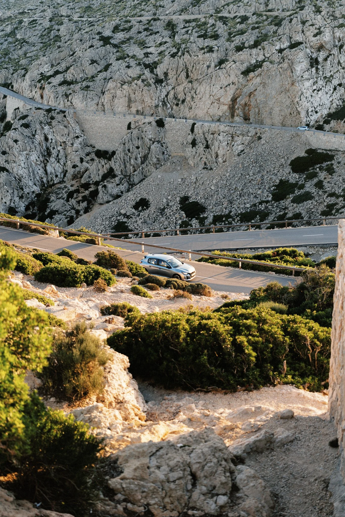 Straße windet sich durch bergige, steinige Landschaft mit grünen Büschen, Autos fahren auf der Straße, hohe Felswände im Hintergrund.