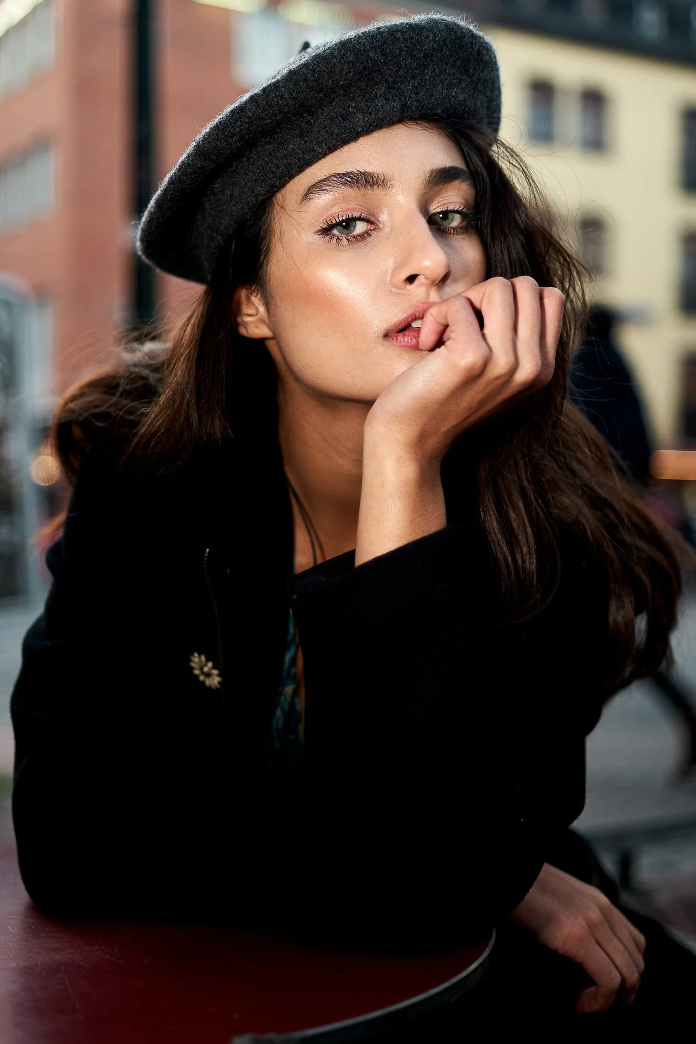 Junge Frau mit dunklen Haaren, grauen Augen und dunkler Kleidung trägt einen schwarzen Wollhut und sitzt an einem Tisch im Freien, während sie nachdenklich schaut.