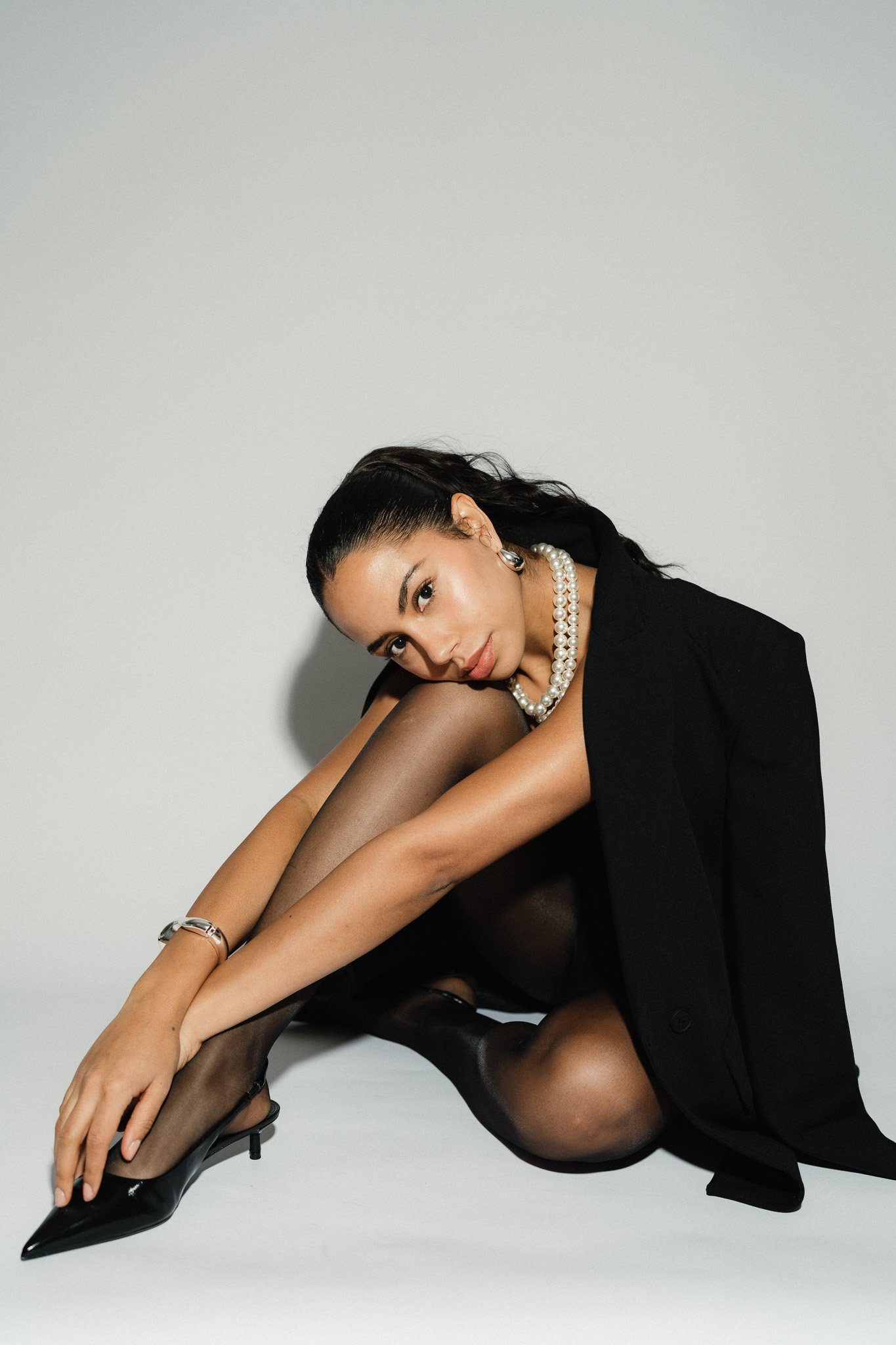 Frau in schwarzem Anzug, schwarzen Nylons, schwarzen High Heels und Perlenkette sitzt vor weißem Hintergrund, mit dunklen Haaren und natürlichem Make-up.