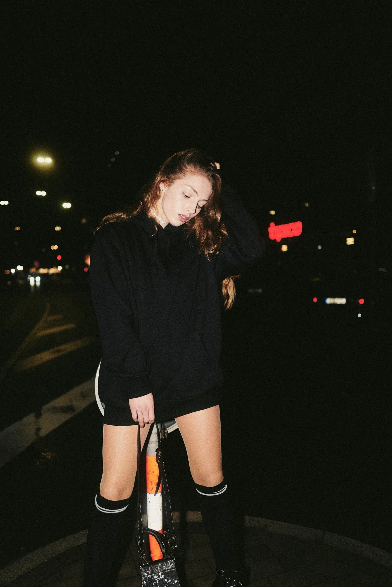 Junge Frau in dunkler Kleidung und hohen Socken steht nachts auf einem Bahnsteig und hält einen Rucksack, die Augen geschlossen, mit einem dunklen Hintergrund mit Straßenlichtern und einem leuchtenden roten Schild im Hintergrund.