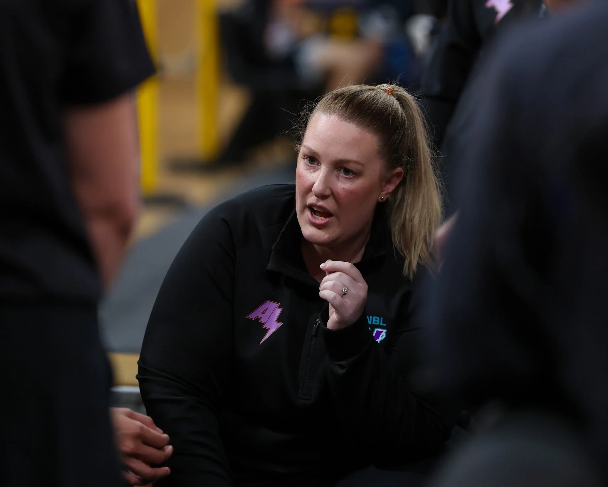 A young woman with blonde hair tied in a ponytail is engaged in a conversation with other people in a group setting. She is wearing a black top and has an expressive facial expression, appearing to speak passionately.
