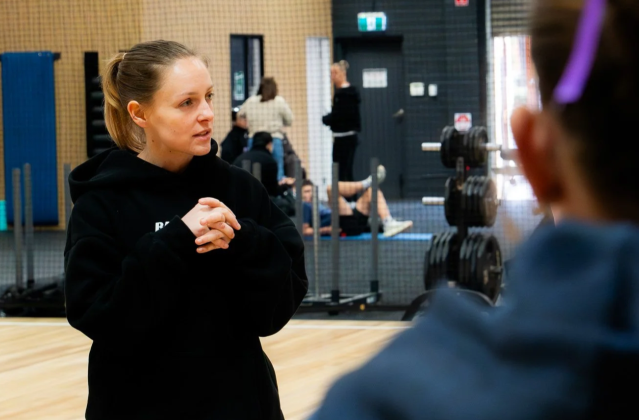 A woman with blonde hair in a ponytail wearing a black hoodie, speaking to another person in a gym with workout equipment in the background.