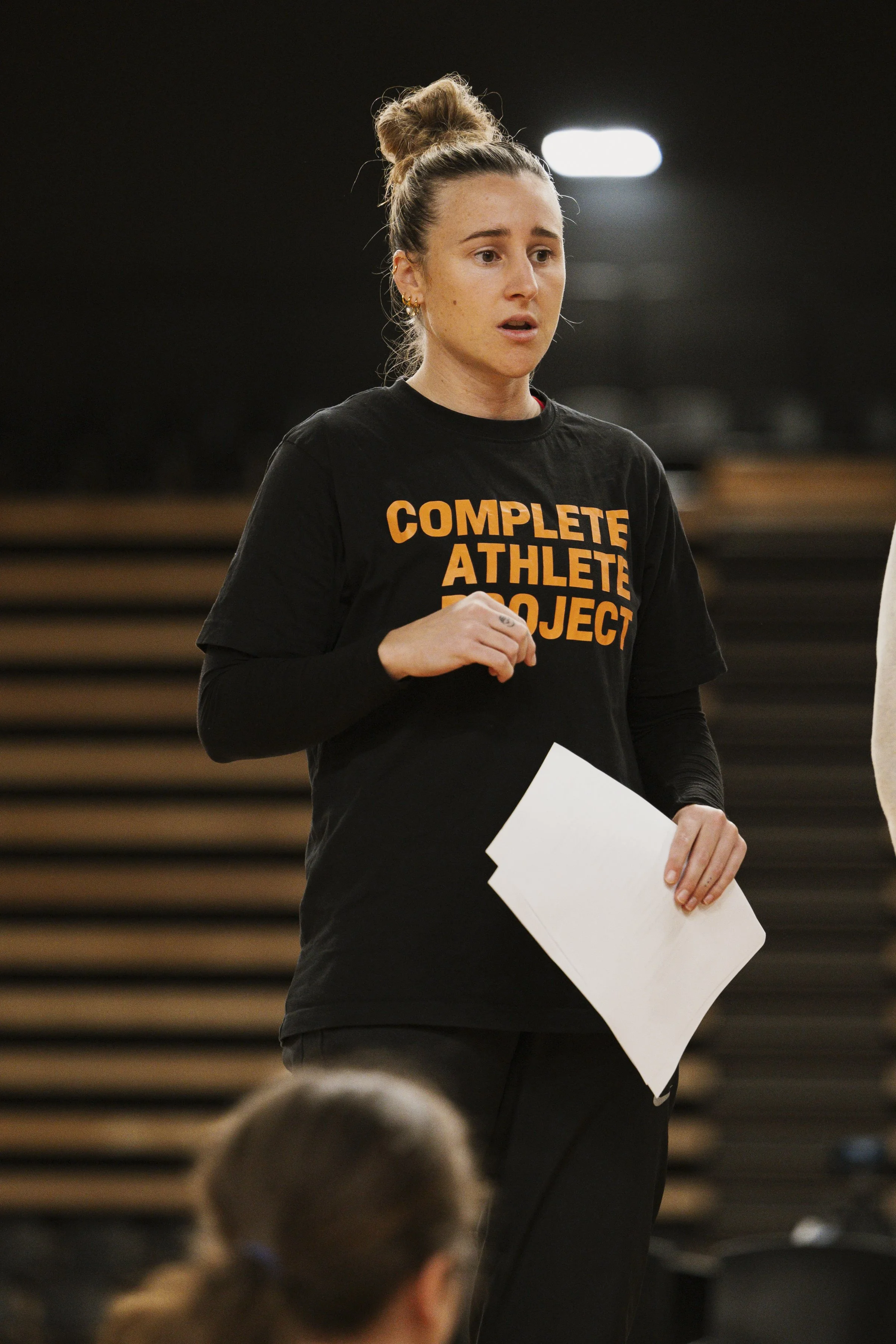A woman with her hair in a bun wearing a black T-shirt that says "Complete Athlete Project" holding papers, standing indoors, possibly in a gym or conference room.