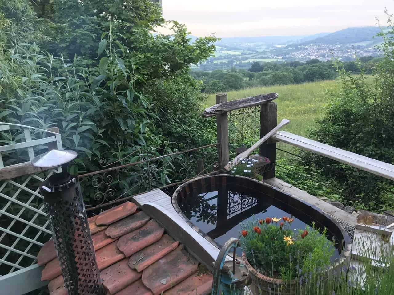 A rustic outdoor tub filled with water, surrounded by lush greenery and flowers, overlooking a scenic landscape with fields, trees, and distant hills.