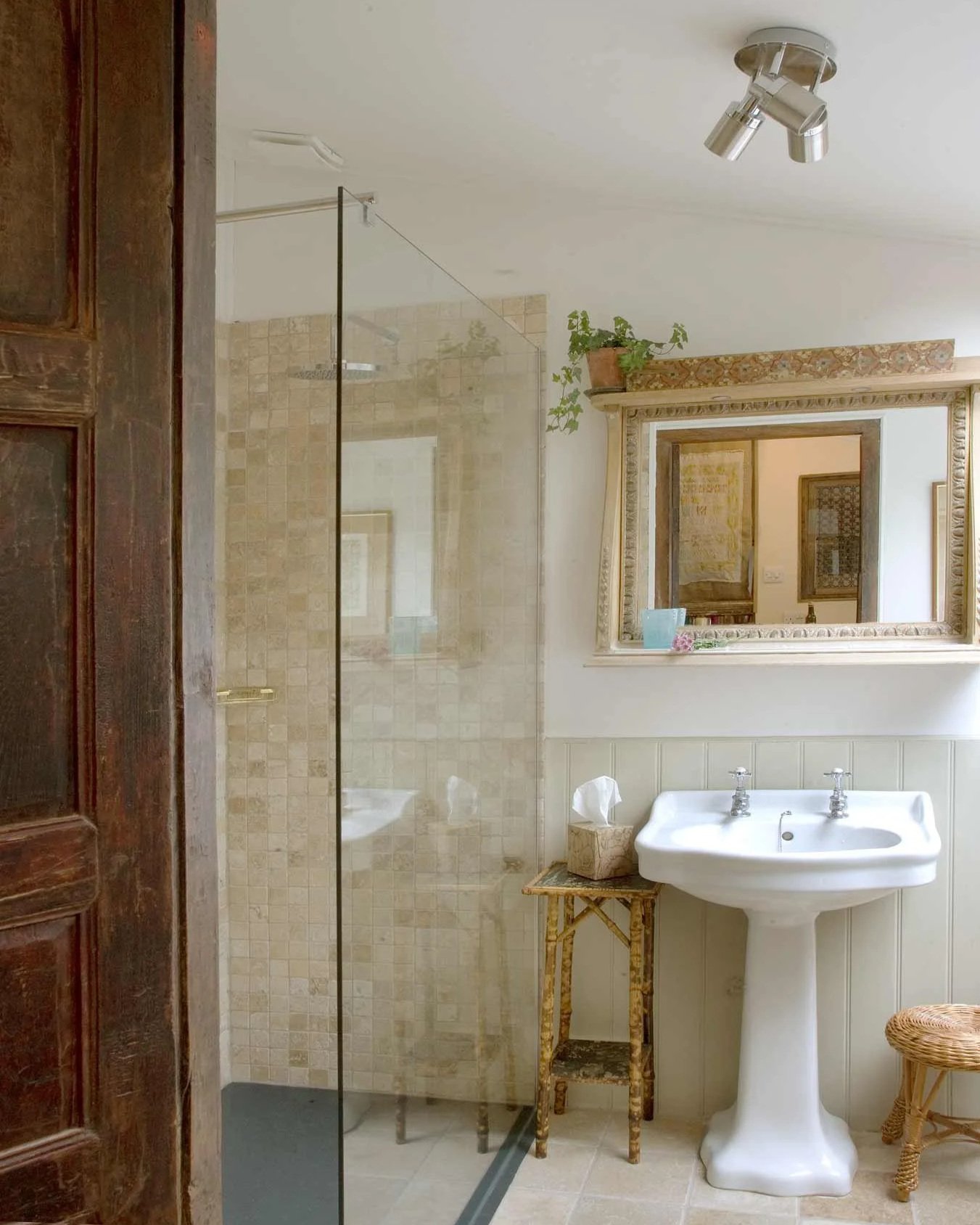 Bathroom with a glass shower, a white pedestal sink, a large decorative mirror, a small table with a tissue box, and wicker stool, with vintage decor and a potted plant on top of the mirror frame.