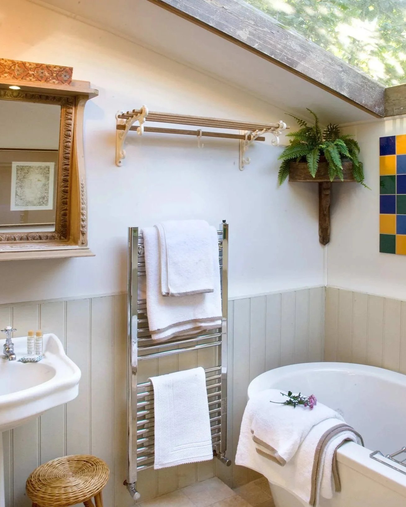 Bathroom with towel rack, bathtub with flowers, small sink, mirror, wicker stool, and potted plant with ferns, under a skylight with sunlight.