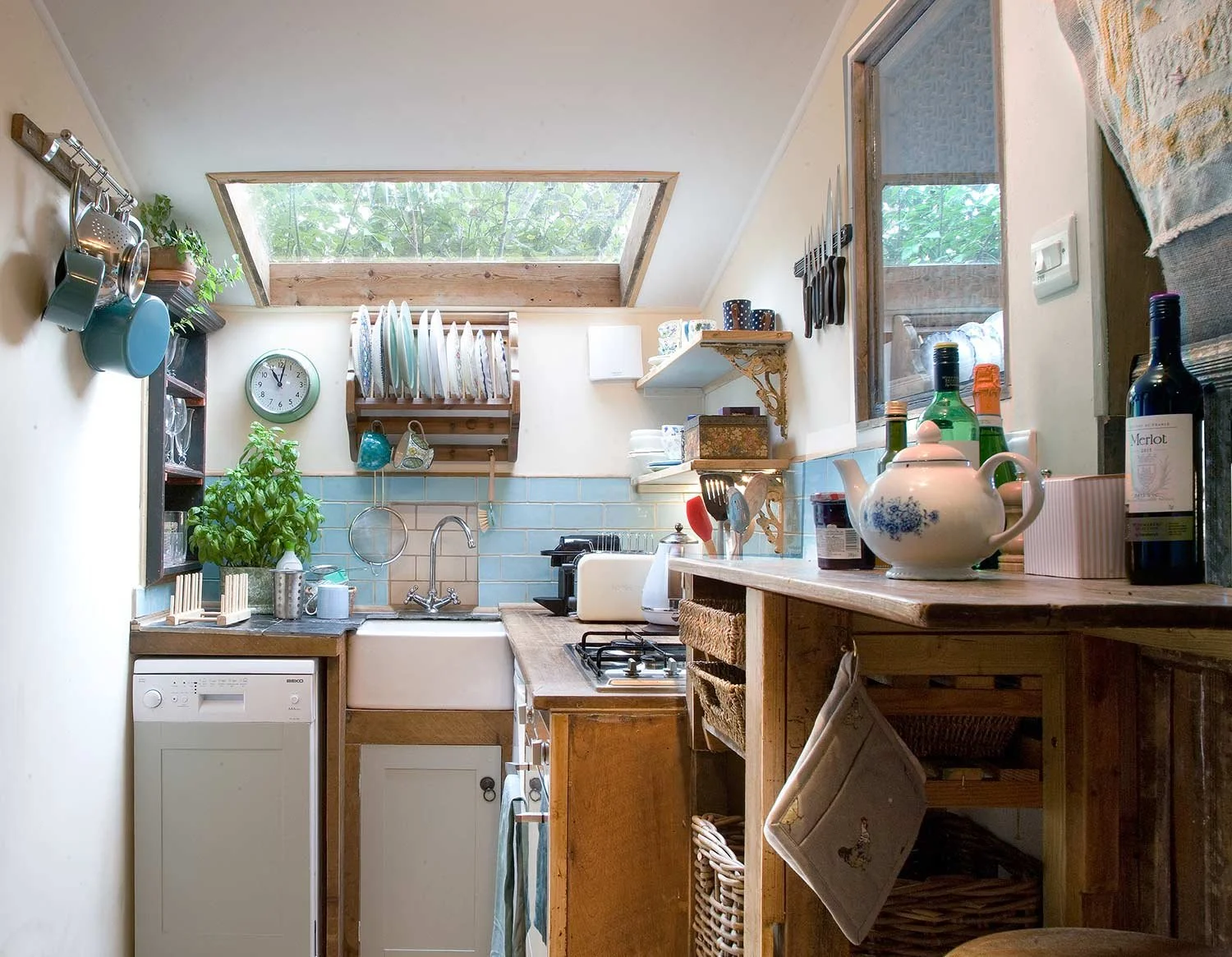 Cozy kitchen with a skylight, wooden countertops, and open shelving with dishes and decorative items, featuring a small washing machine beside the sink, potted plant, and various kitchen utensils and bottles on the counter.