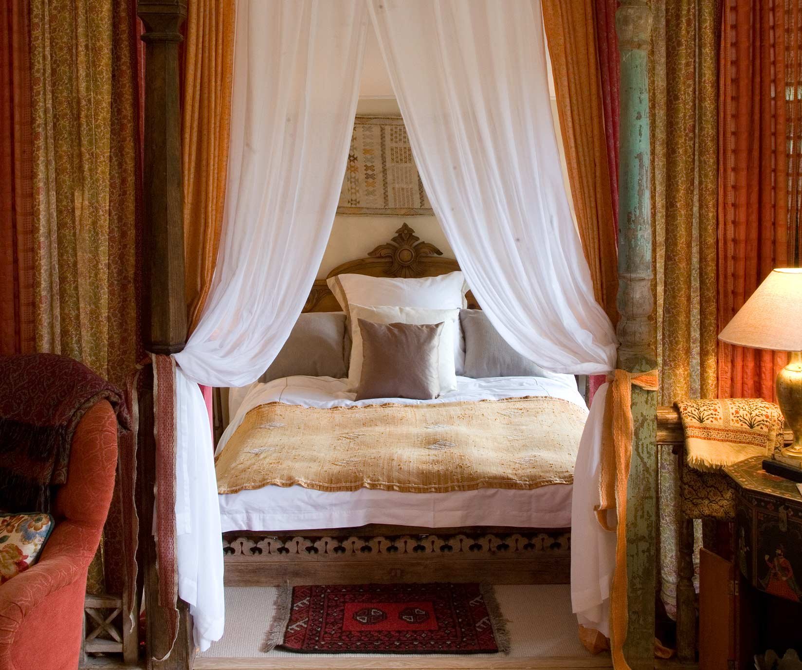 A four-post bed with white canopy and beige bedding, with decorative pillows. There is a lamp on a table to the right, and a patterned rug on the floor. Curtains and wall decor frame the bed.
