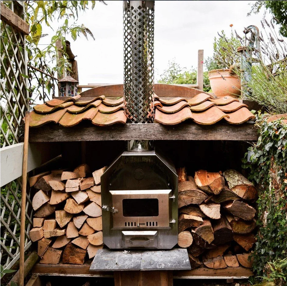 A wood-fired outdoor oven with a metal door, stacked firewood on both sides, and a sloped roof with red clay tiles, situated in a garden with plants and decorative items.
