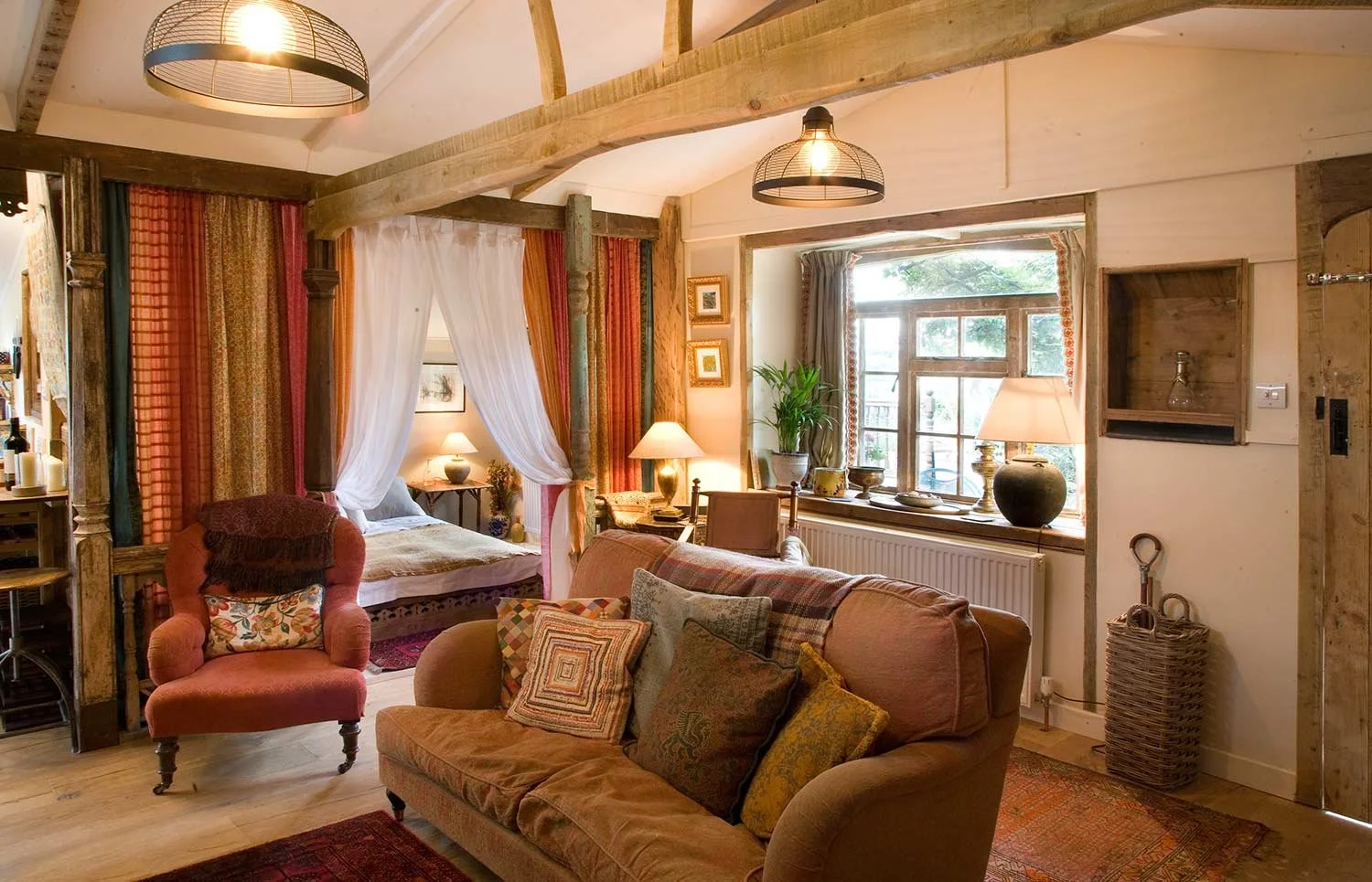 A cozy living room with a bed area separated by a canopy of sheer and patterned curtains. There's a pink armchair with a floral pillow, a peach-colored sofa with multiple decorative pillows, a wooden side table with lamps, a window with curtains and potted plants, and warm lighting creating a rustic, inviting atmosphere.