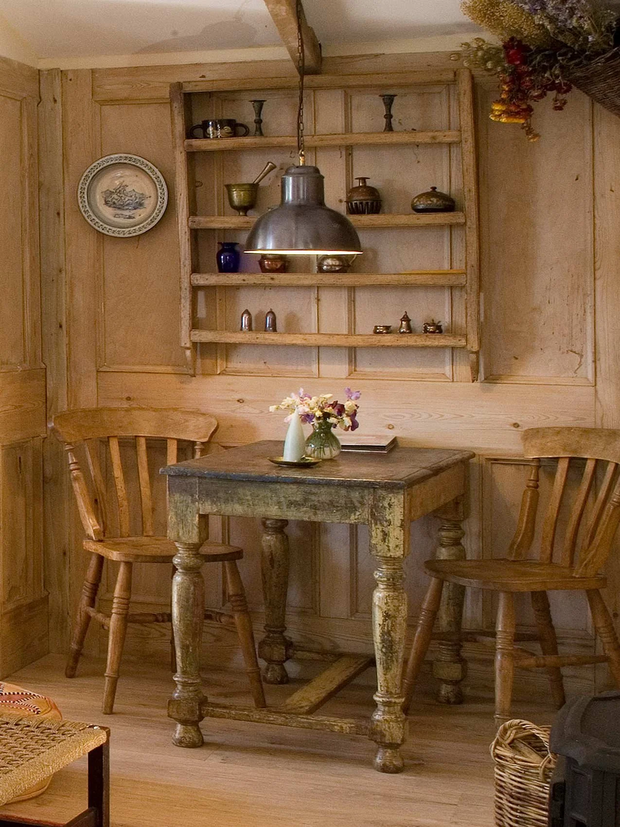 A cozy corner with a rustic wooden table and two matching wooden chairs, a flower vase with pink and purple flowers, wooden wall paneling, and a wall-mounted wooden shelf with decorative pottery and a plate.