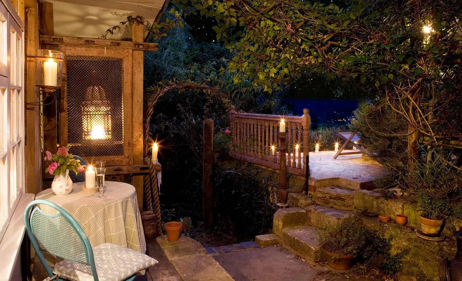 An outdoor backyard patio at night with lit candles, potted plants, a small table with a floral vase and candles, a wooden fence, and a view of a lit cityscape in the distance.