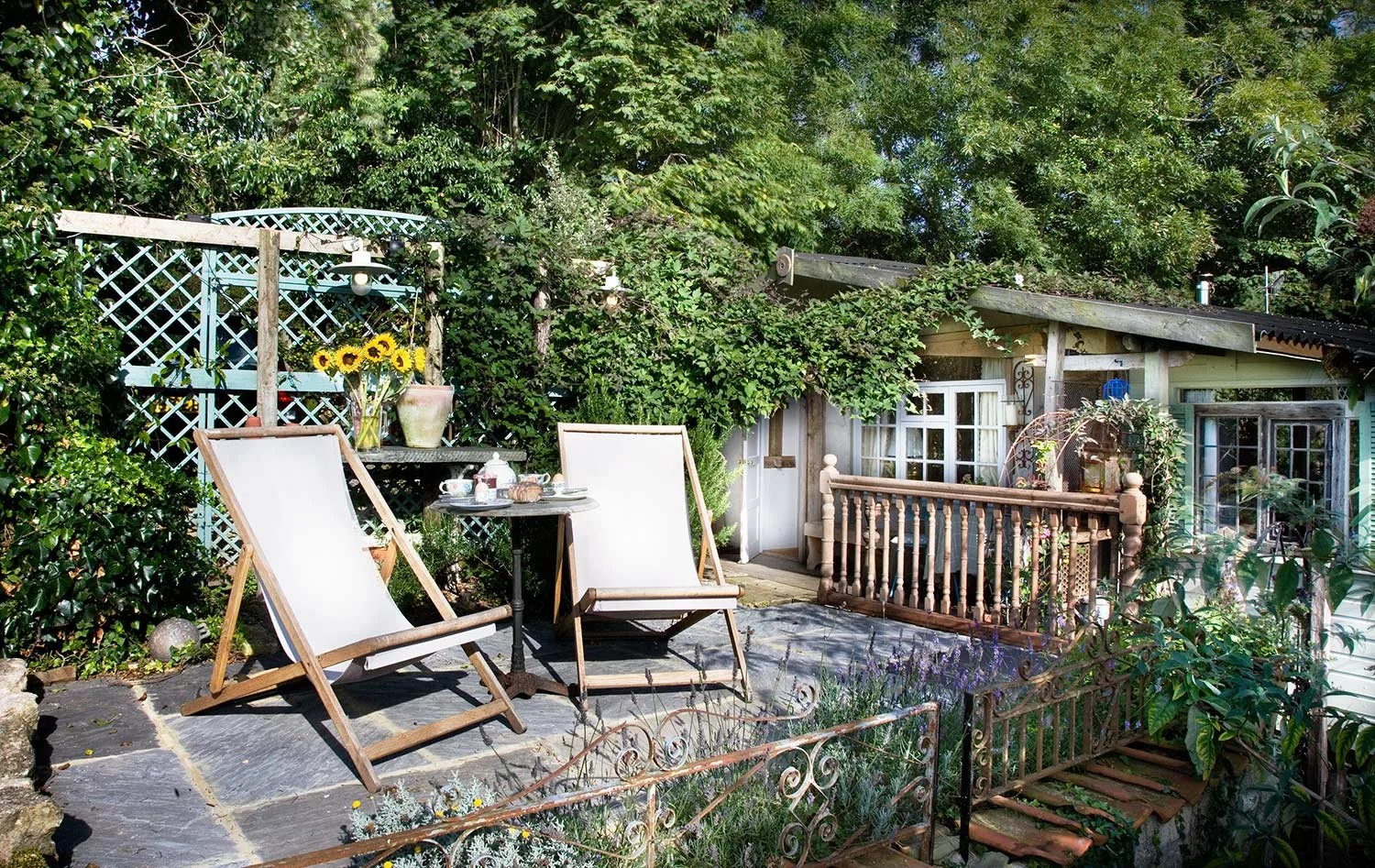 A cozy outdoor patio area with two wooden deck chairs, a small round table with tea cups, a vase of sunflowers, and a small garden shed surrounded by lush green trees and plants.