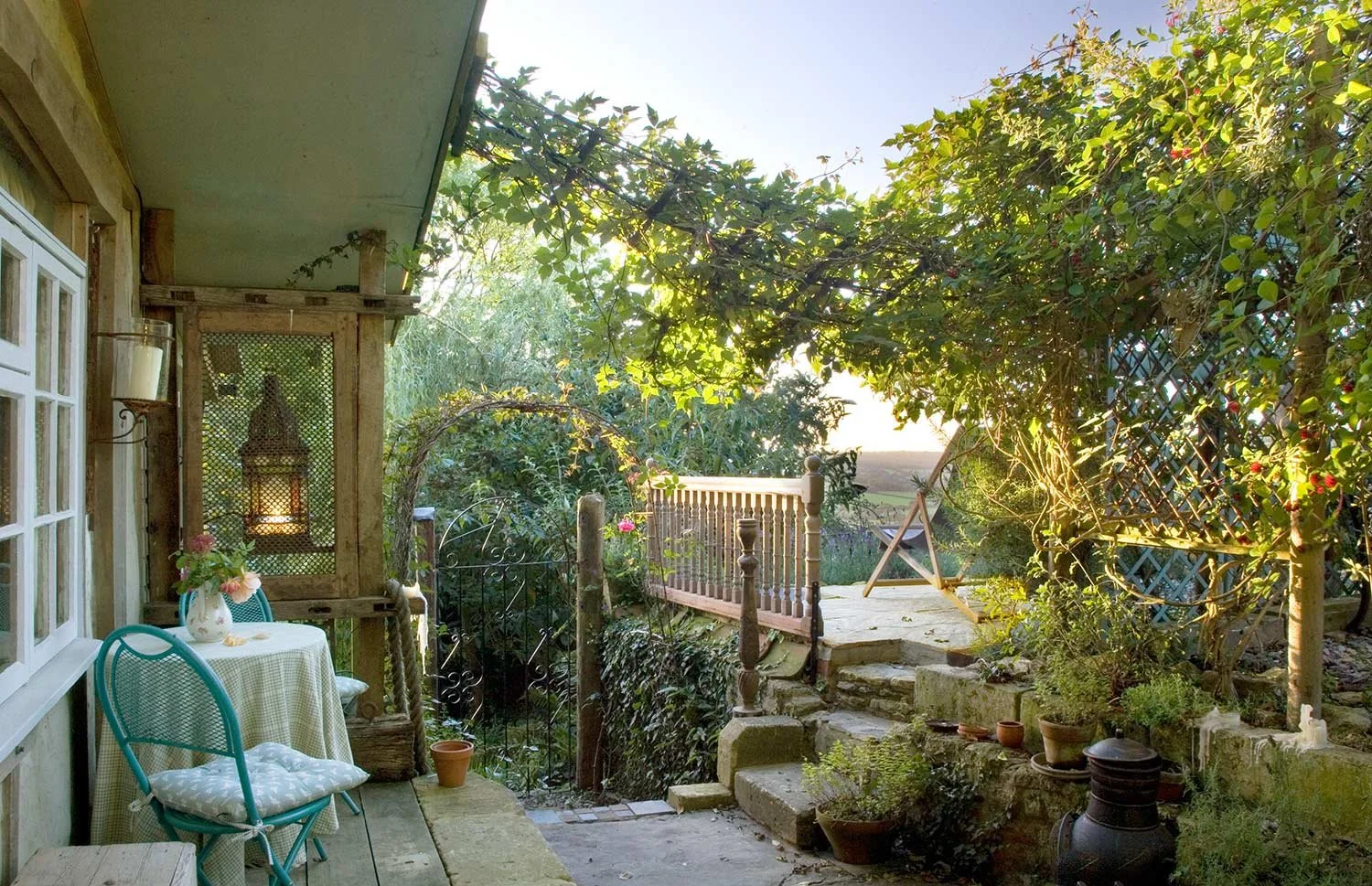 A cozy outdoor patio area in the late afternoon sunlight, with a small round table covered with a checkered tablecloth, a flower vase, and two metal chairs, surrounded by potted plants, a wooden railing, and lush greenery.