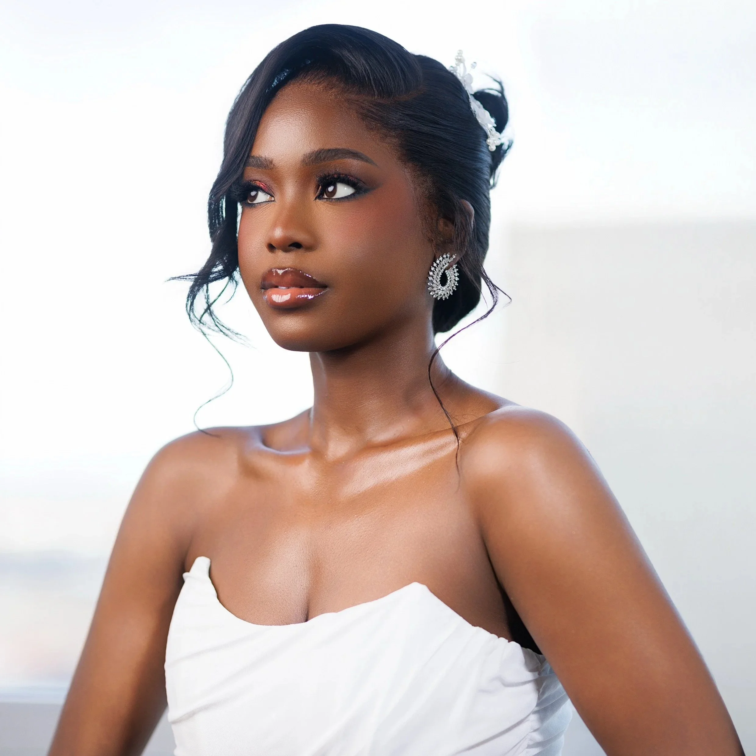 A woman with styled black hair, wearing diamond earrings, in a white strapless dress, posing against a blurred light background.