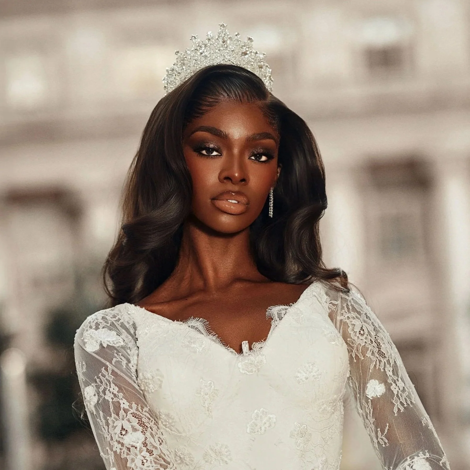 A woman with dark skin, long wavy hair, and wearing a crown and white lace dress stands confidently in a grand hallway.