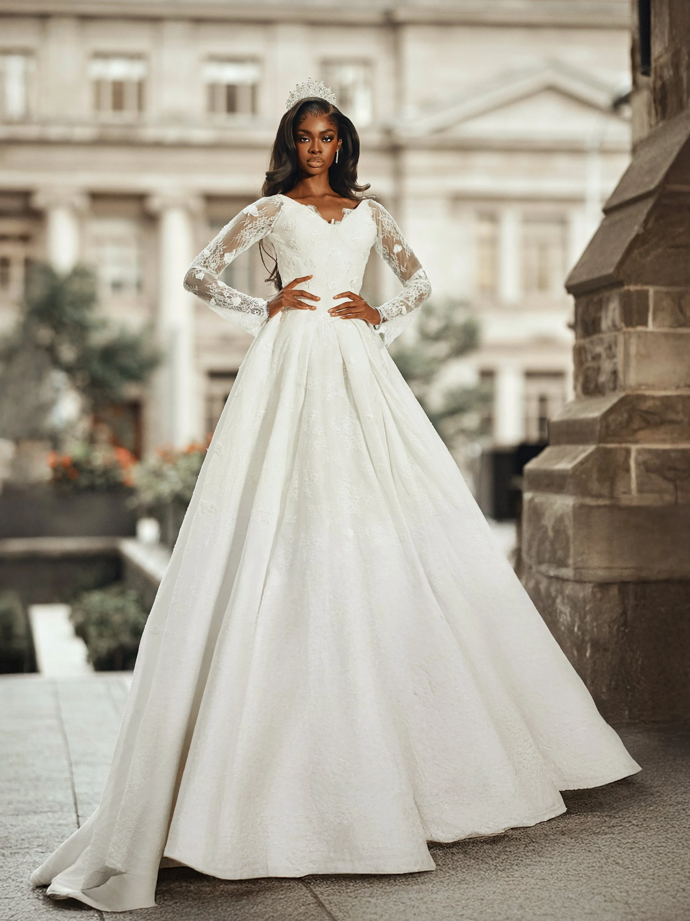 A woman in a white wedding dress with lace sleeves and a tiara, standing outdoors in front of a building with classical architecture.