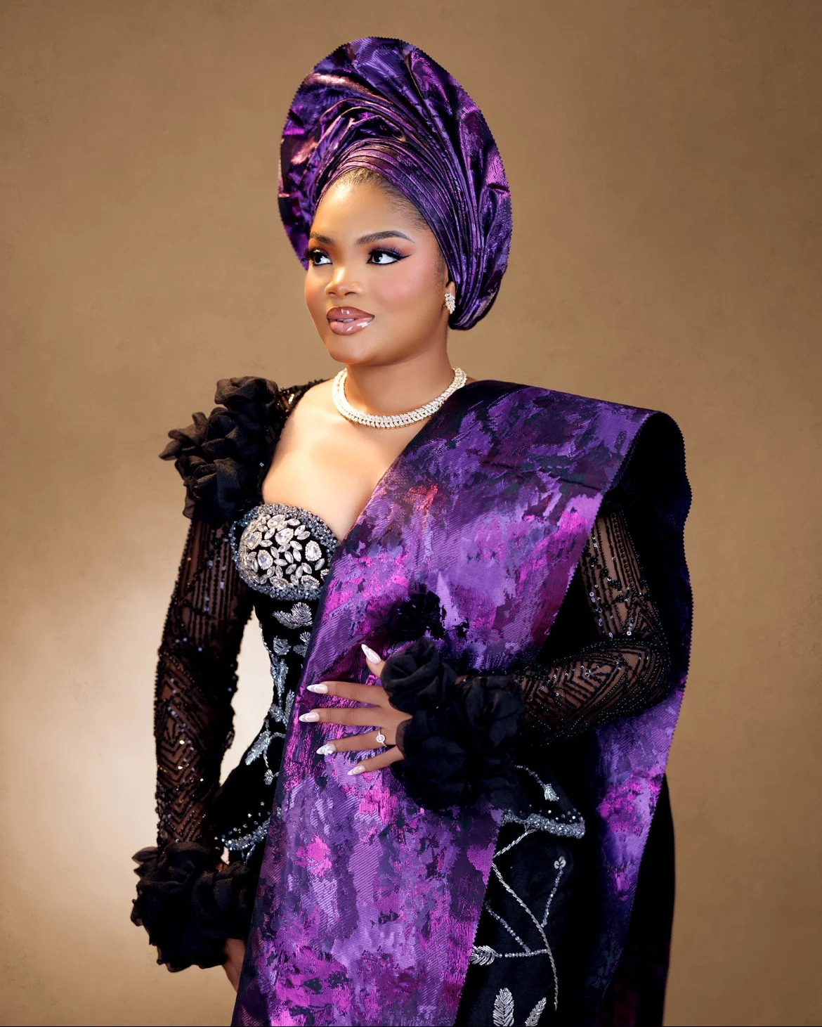 A woman in traditional Nigerian attire wearing a purple gele headwrap and a black and purple agbada with intricate embroidery, accessorized with a pearl necklace and diamond jewelry.