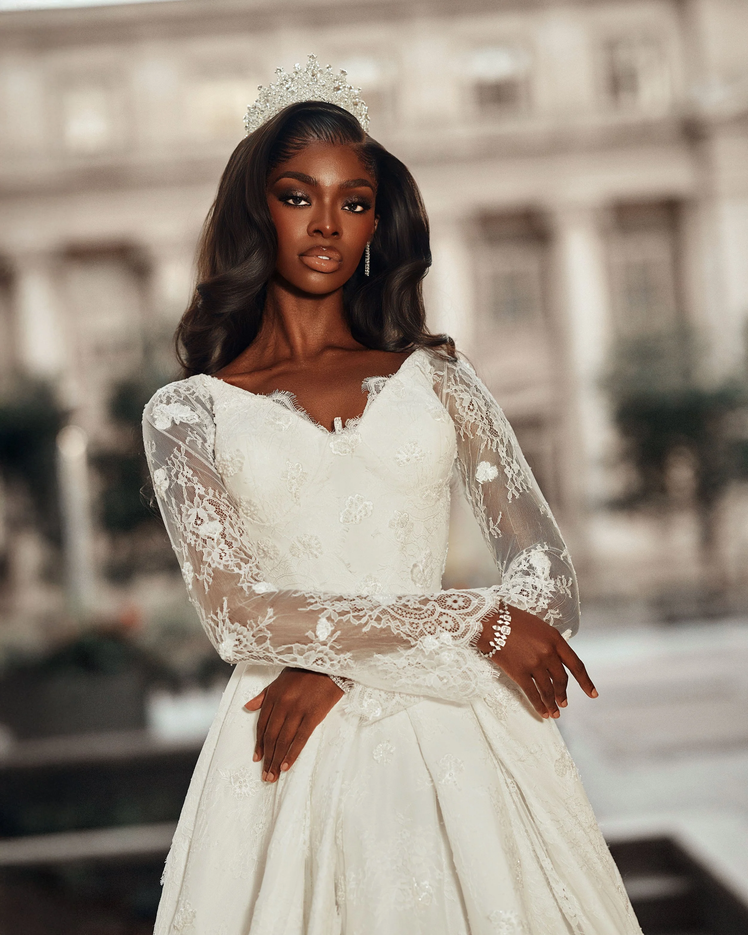 A woman in a white lace wedding dress with a tiara stands outdoors, with a blurred cityscape background.