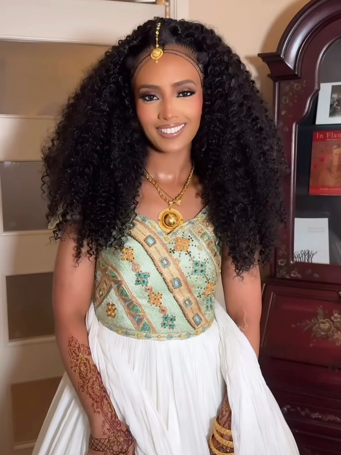 A young woman with long curly black hair, gold jewelry, and henna arm art, wearing a traditional embroidered outfit, standing indoors.