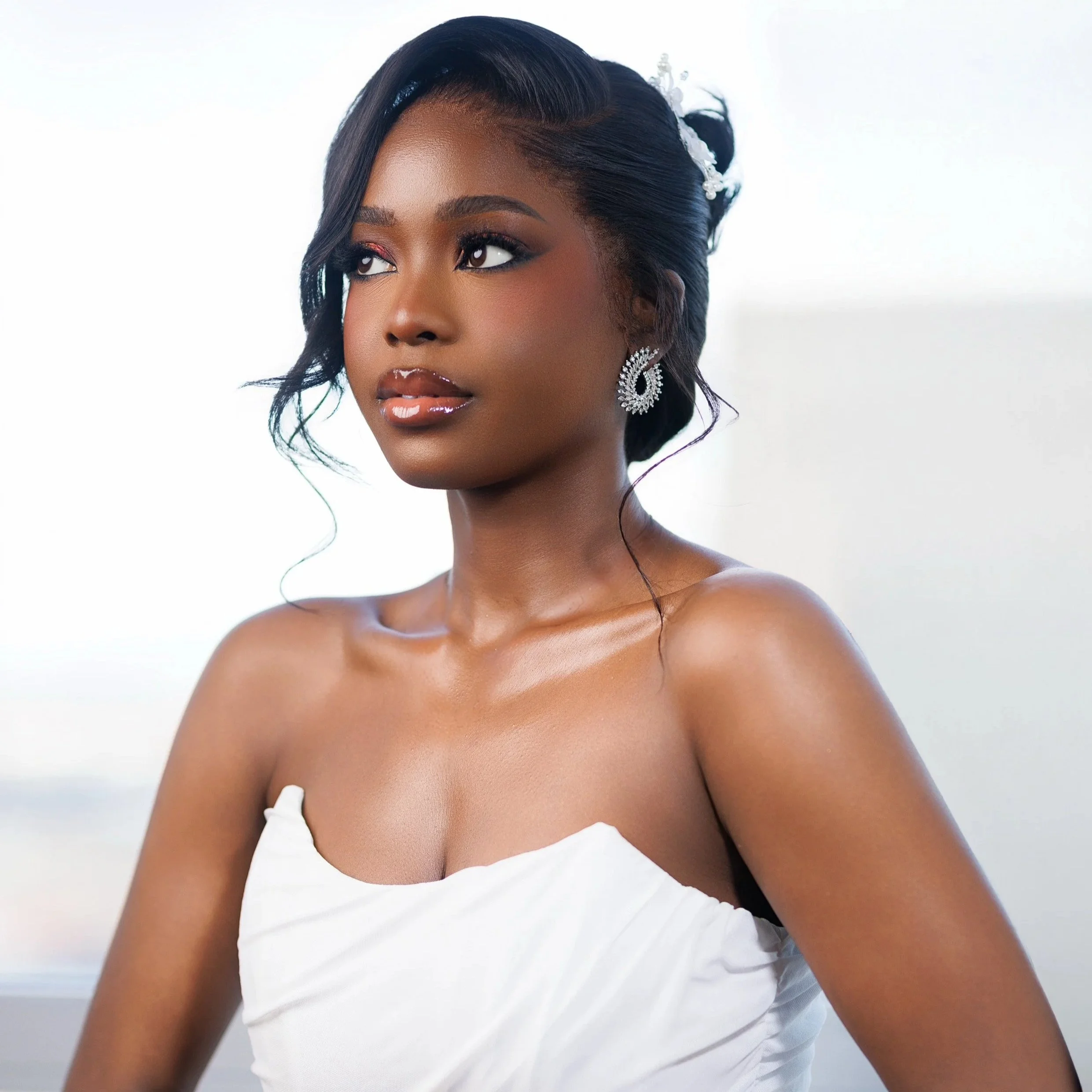 Portrait of an elegant woman with dark hair styled in waves, wearing sparkling earrings, a strapless white dress, and glossy lips, with a blurred white background.