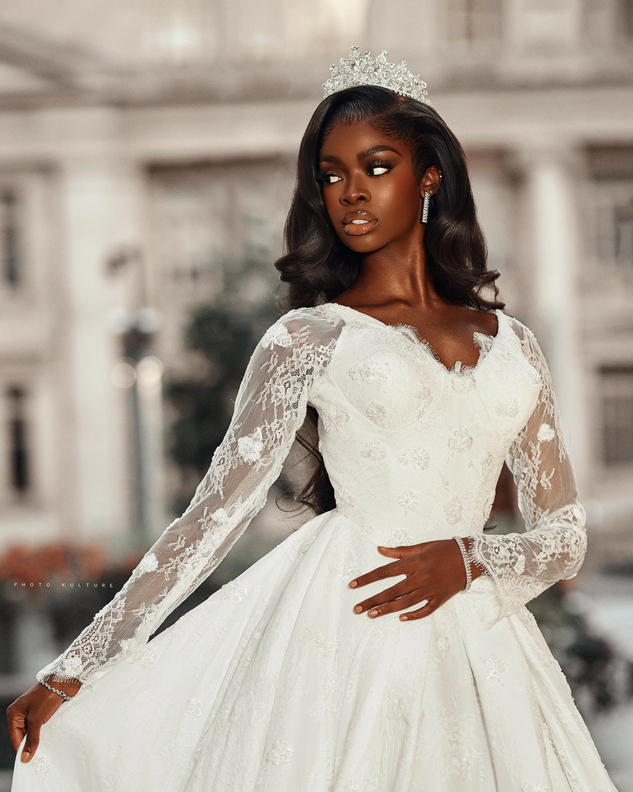 A woman in a white wedding dress with lace long sleeves and a full skirt, wearing a tiara and earrings, with dark wavy hair.