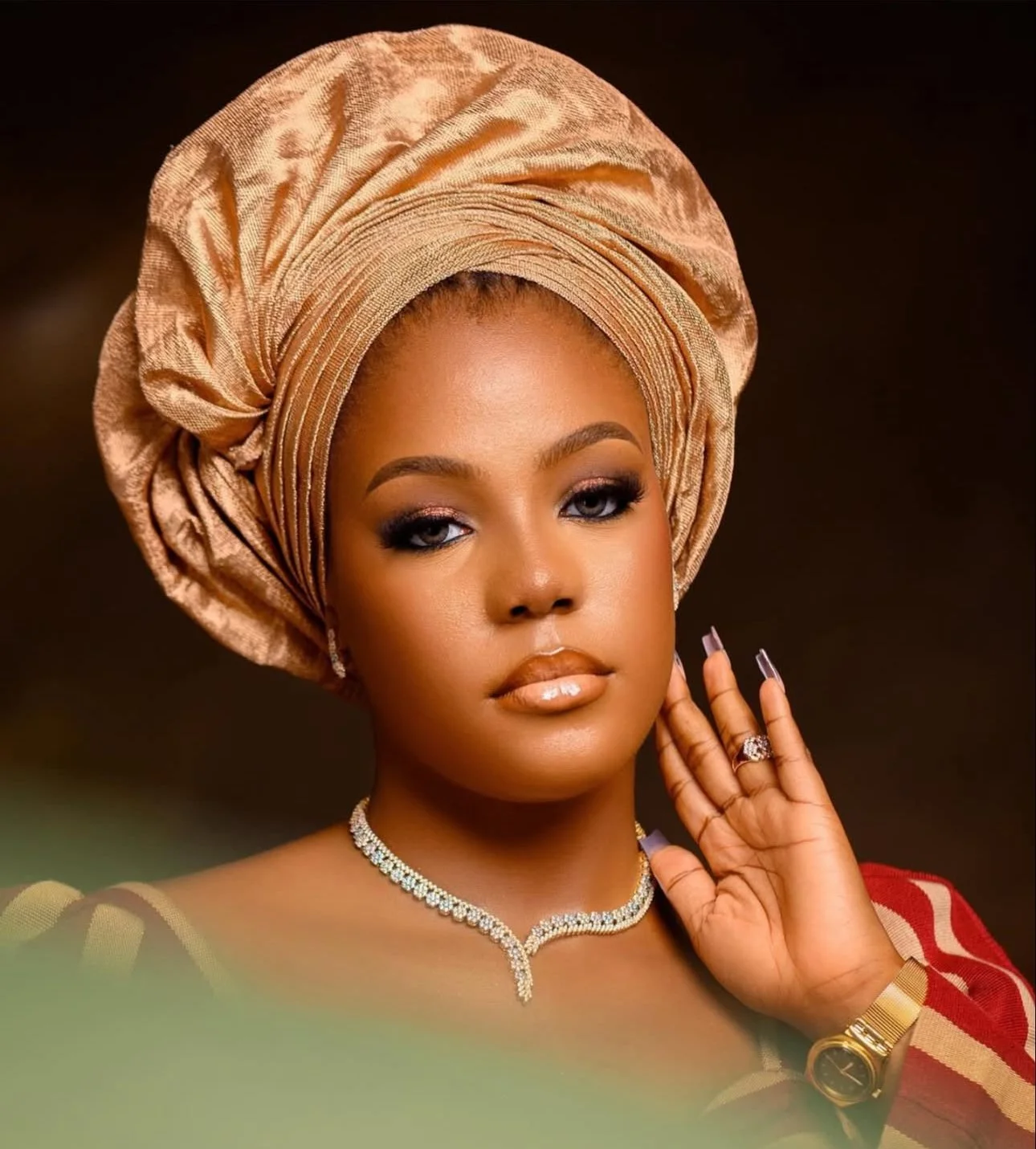 A woman wearing a gold headwrap, diamond jewelry, a watch, and a red and beige outfit, posing with her hand near her face.