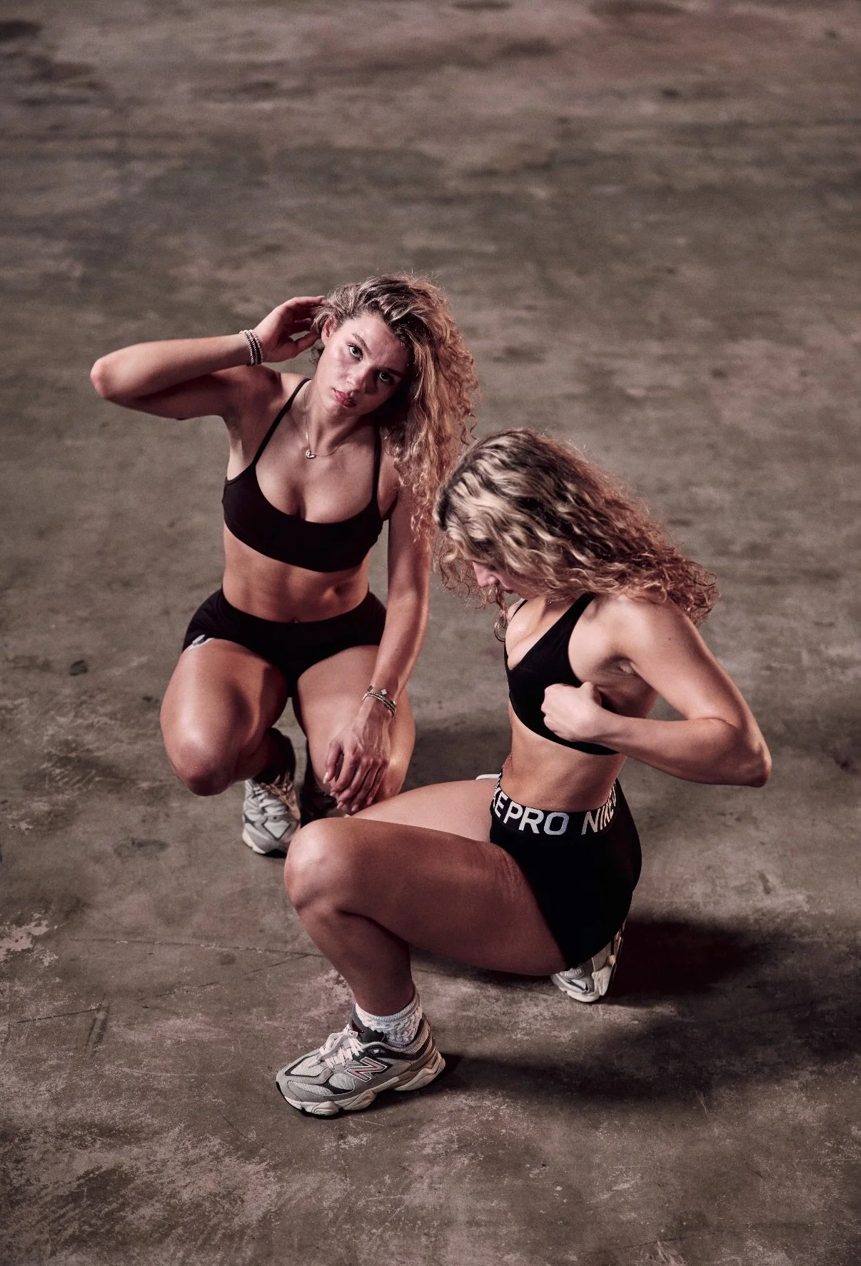 Two women with curly hair are posing on a concrete floor. One woman is kneeling with her arm flexed, and the other woman is crouching with her hand on her knee, looking at the camera. They are wearing athletic clothing.