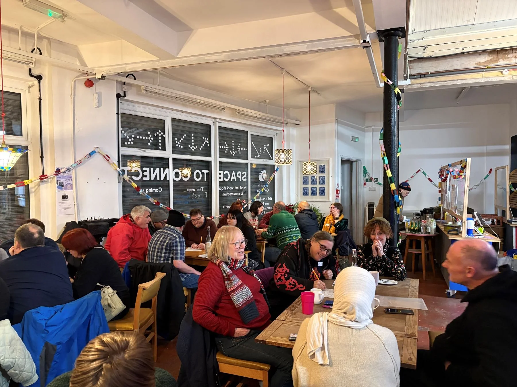 Groups of people of all ethnicities and ages sit around tables. They have paper and pens and some are writing. They are in a white room with colourful paper chains swagged along the walls.