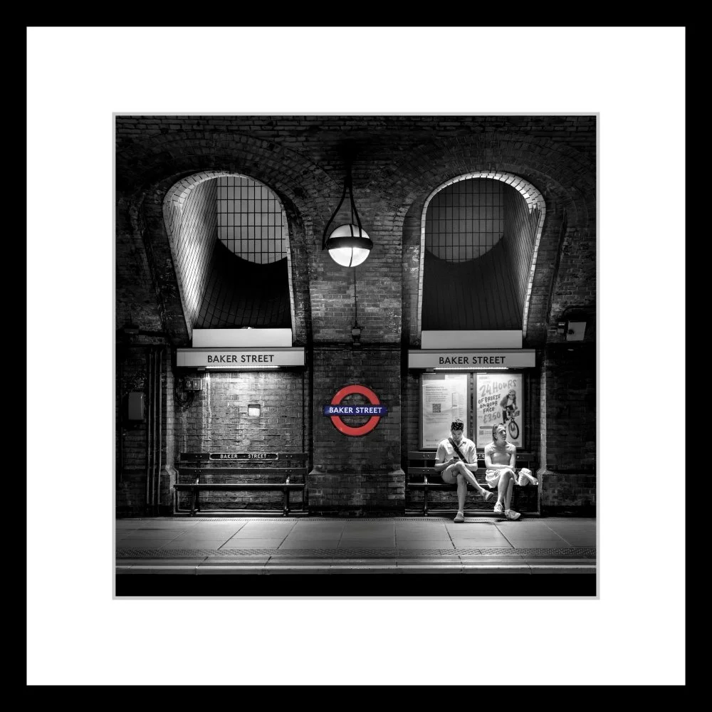 Black‑and‑white photograph of Baker Street Underground Station, showing two people seated on a bench beneath arched brickwork and illuminated signage.