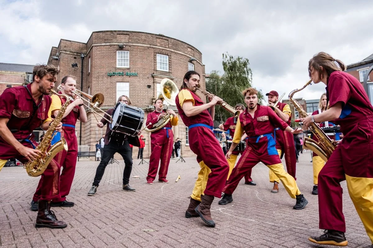 The image is outside in a city centre. A group of musicians wearing red and yellow boiler suits are performing.