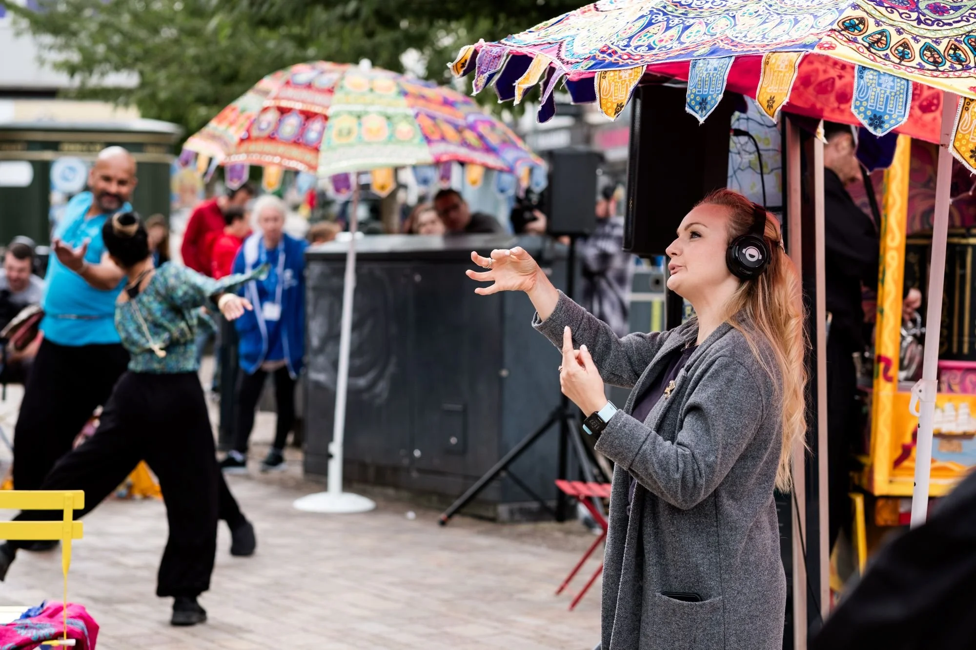 A lady wearing headphones is standing in the foreground side-on to us. Her hands are animabted as she speak in British Sign Language. There are tall umbrellas made with sari fabrics and two people dance in the background.