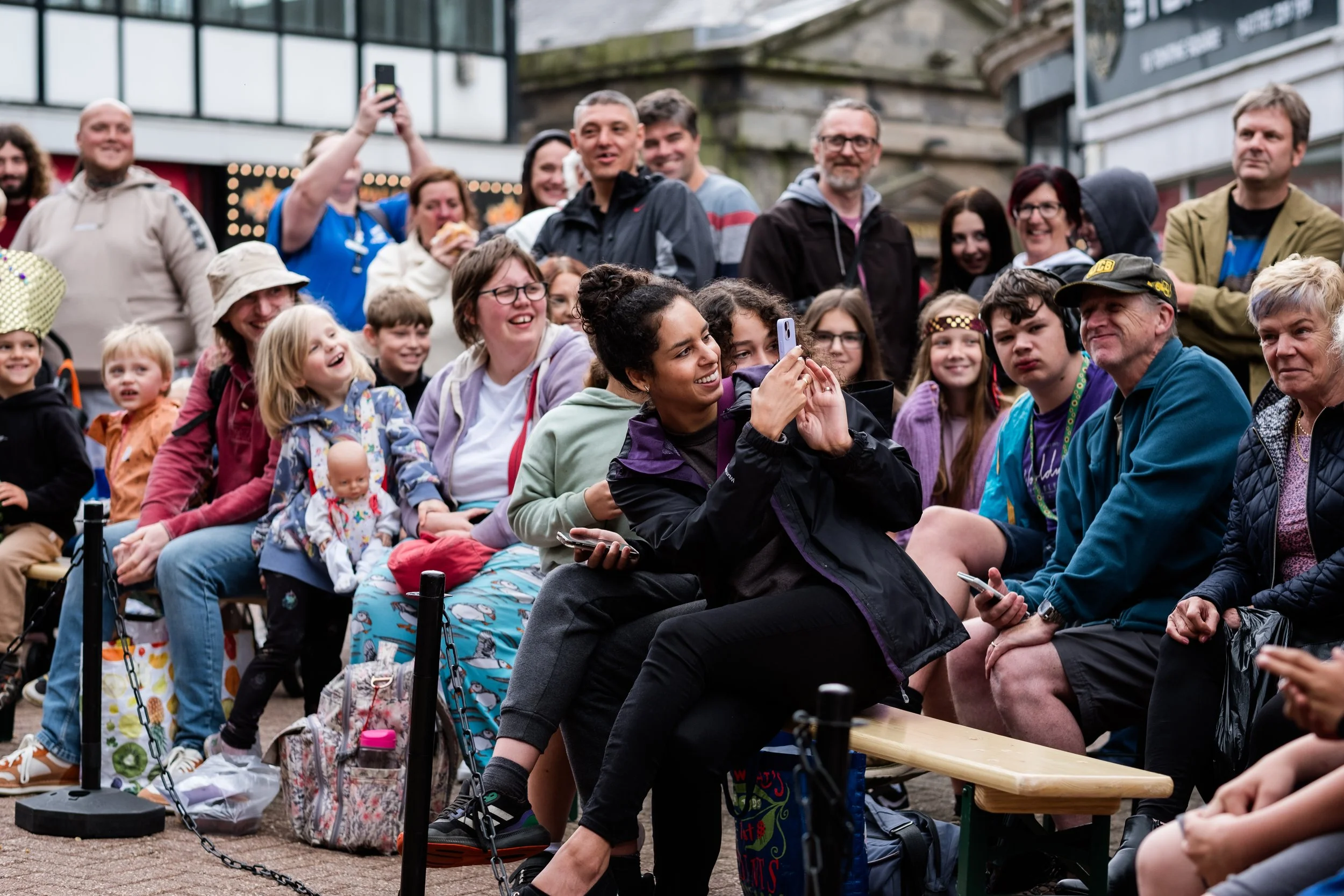 A number of people are sitting down or standing up, smiling,  as though watching a performance. Everyone is looking to the right and a lady in the centre of the image is holding her phone up to take a photograph.