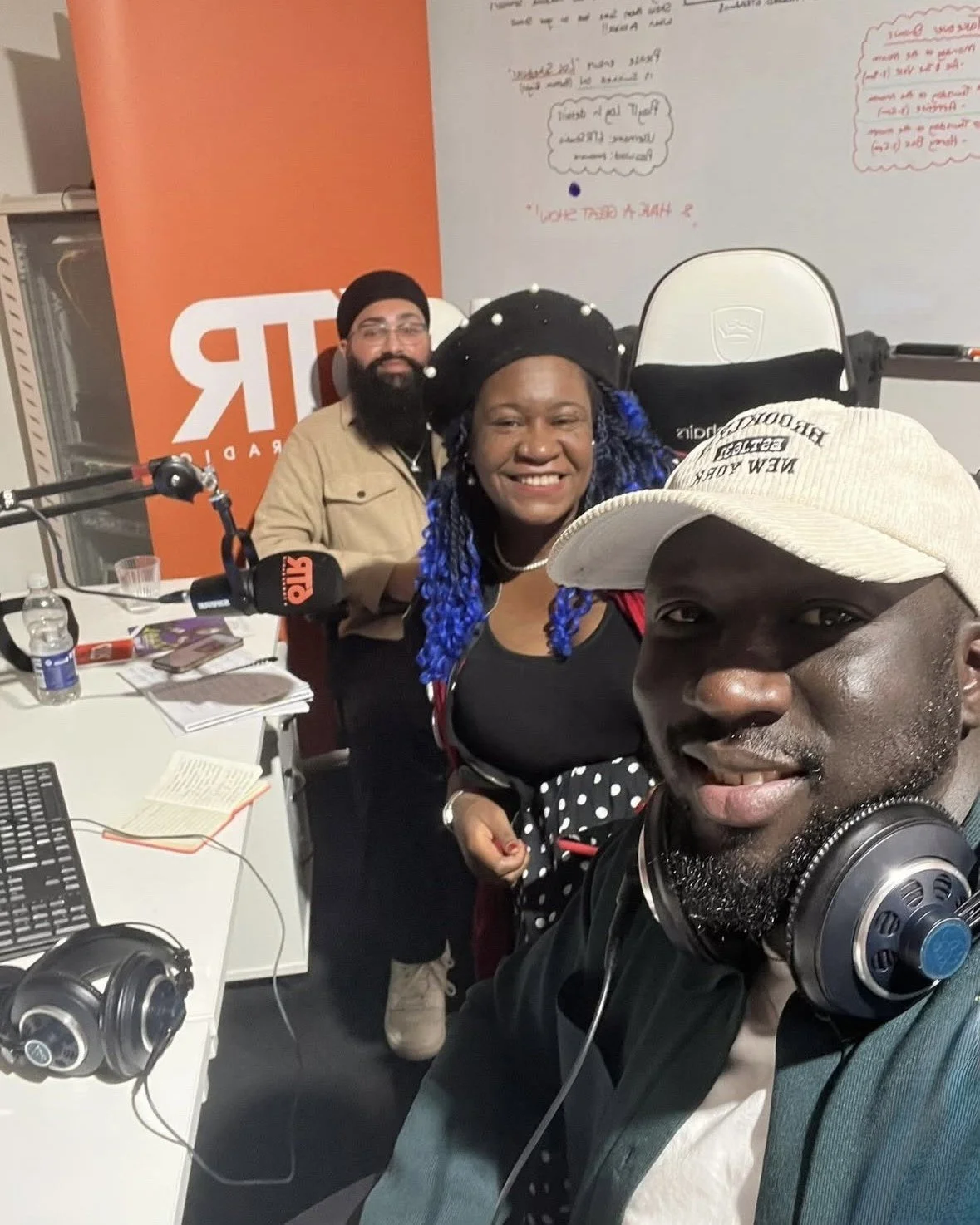 A Black woman with a white cap in the foreground smiles at the camera with headphones around his neck. A Black woman with a black beret is in the middle smiling at the camera and an Asian man with long black beard is in the background.