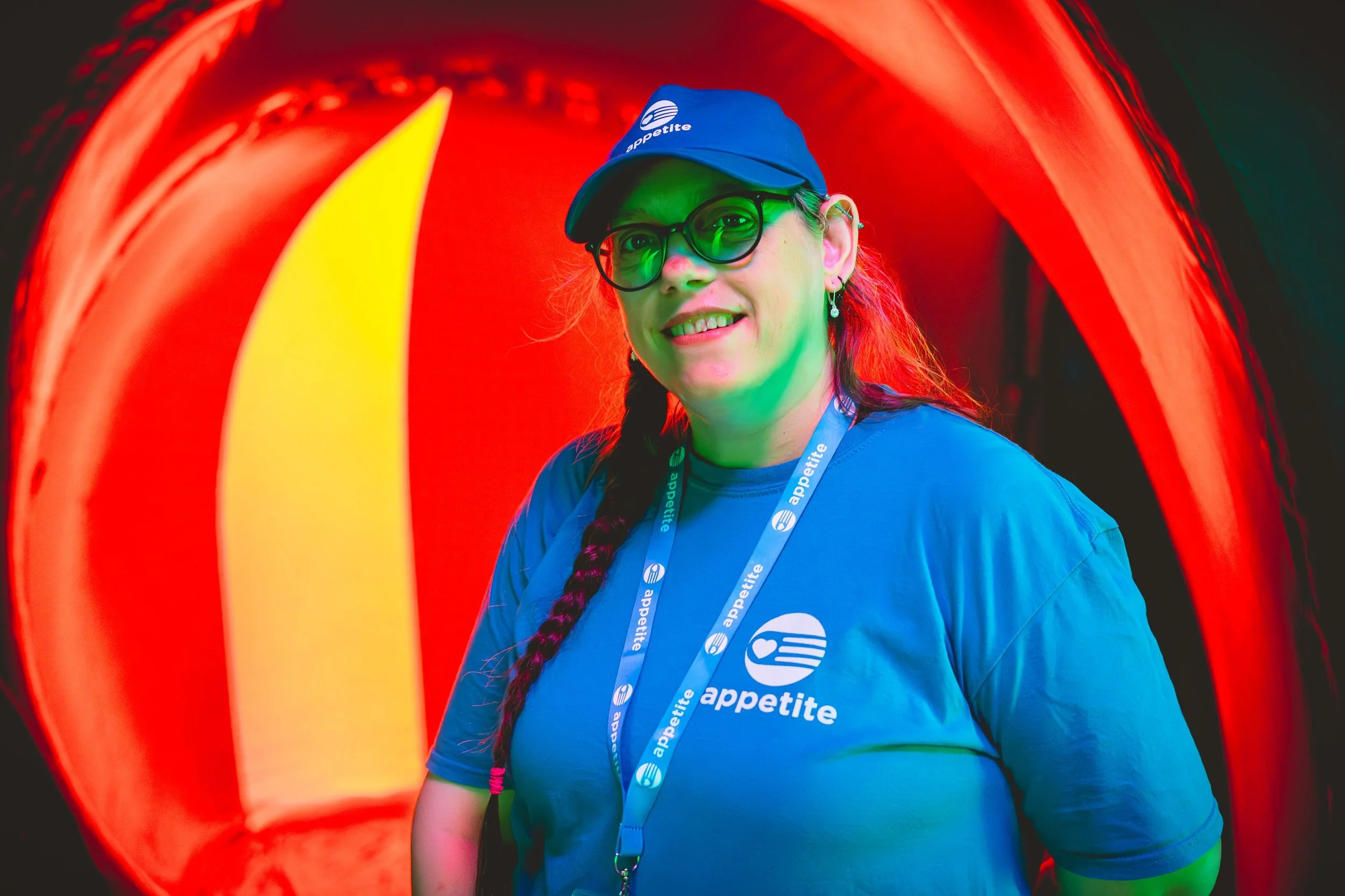 Appetite volunteer Jackie is looking at us with a friendly smile. She has glasses and a long, brown plait over her right shoulder. She's all in Appetite blue with a branded baseball cap. Jackie is surrounded by glowing reds and yellows.