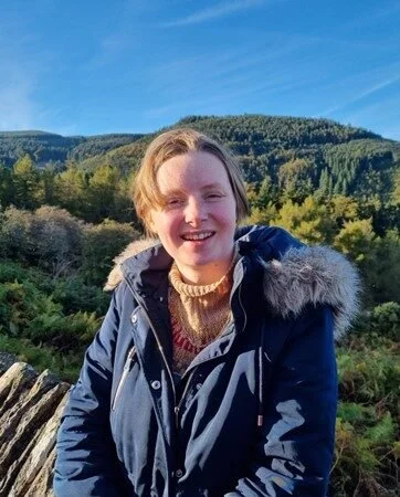 Emma is smiling straight at us from high up in a beautiful countryside scene on a clear, sunny day. They are wearing a roll-neck jumper and dark blue coat with a fur-trim hood.