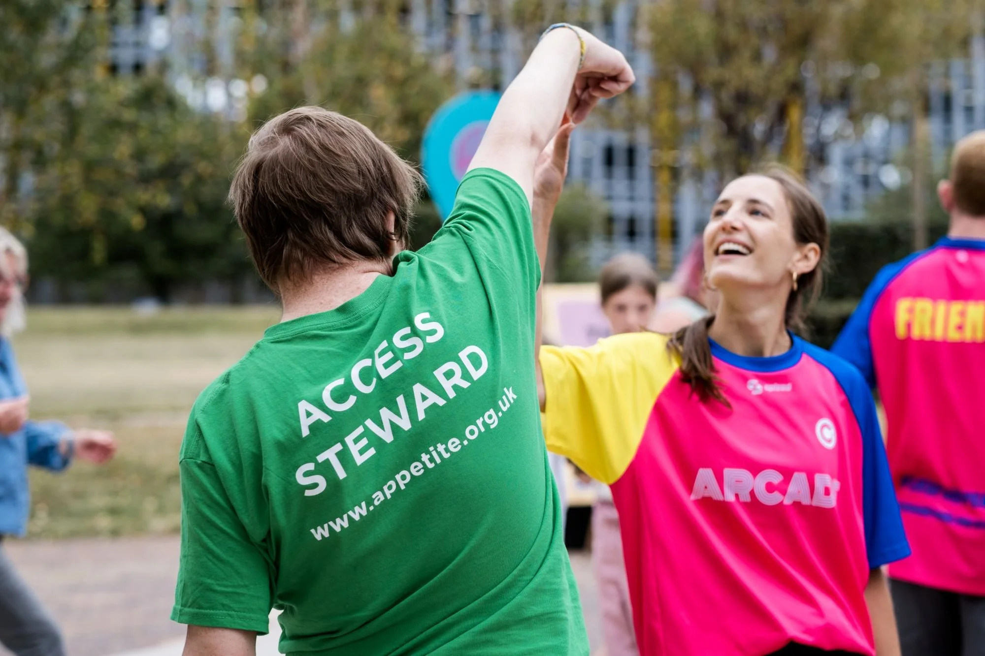 Two people in bright-coloured T-shirts are facing each other, joining right hands in the air. The figure with their back to us wears a green T with Access Steward written on.