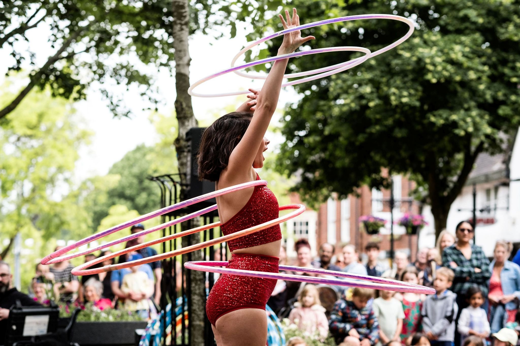 A woman with shoulder length dark hair pictured from the side spinning 5 colourful hula hoops, wearing a red sparkly top and shorts outfit. An audience watches in the background.
