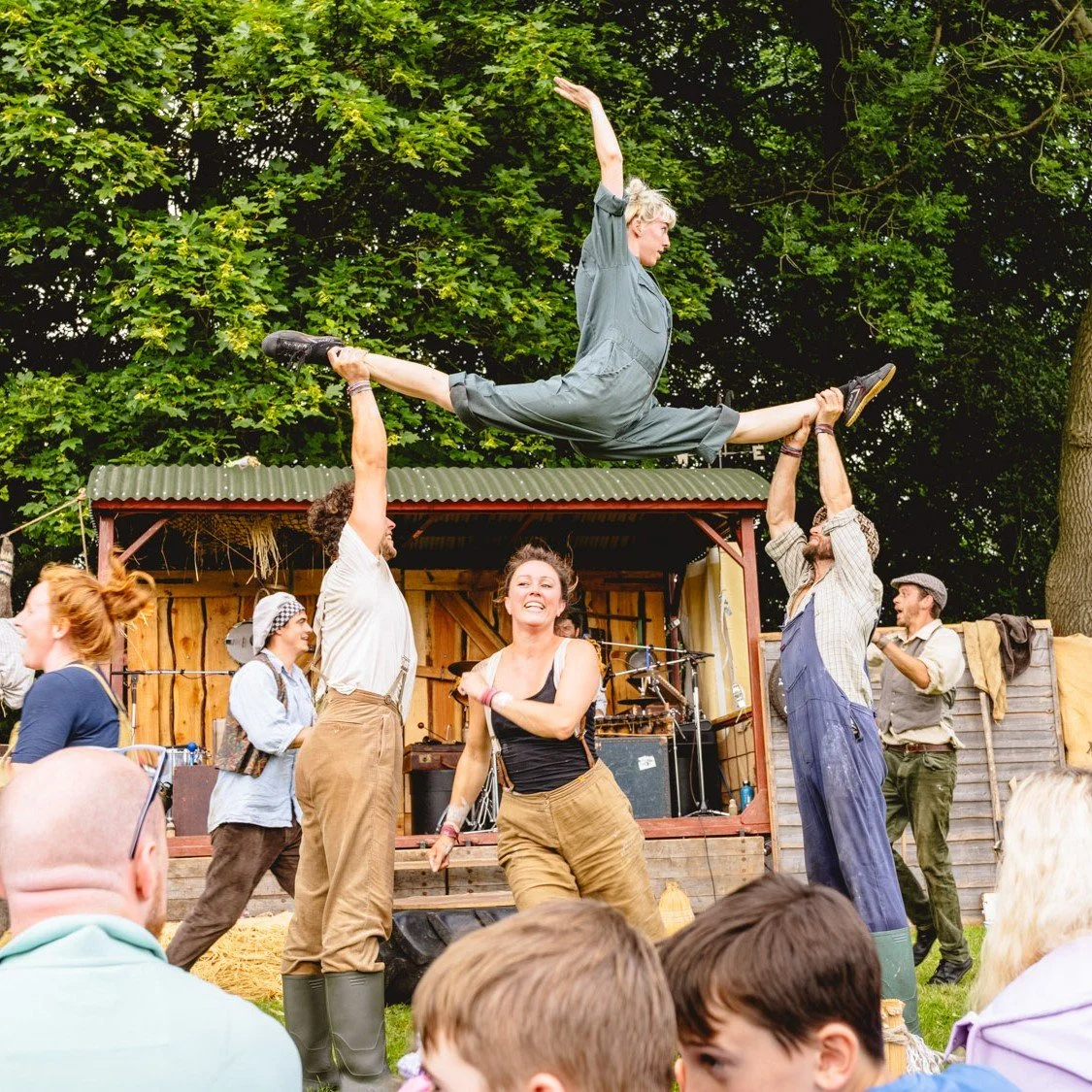 Circus performers dressed in farmer attire (corduroy trousers, shirts, caps and wellies) perform a show. A woman is doing the splits in the air supported by 2 men holding her feet. Another woman runds underneath.