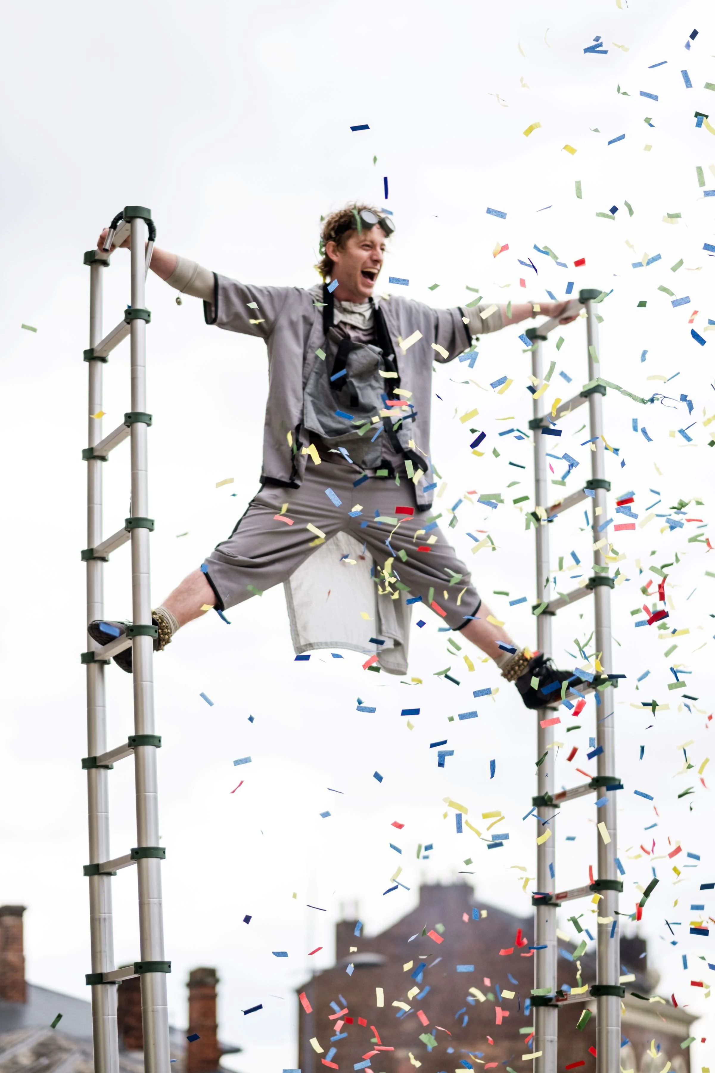 A white man in a grey shirt and shorts stands with arms and legs stretched between 2 ladders, surrounded by colourful confetti and with an excited expression