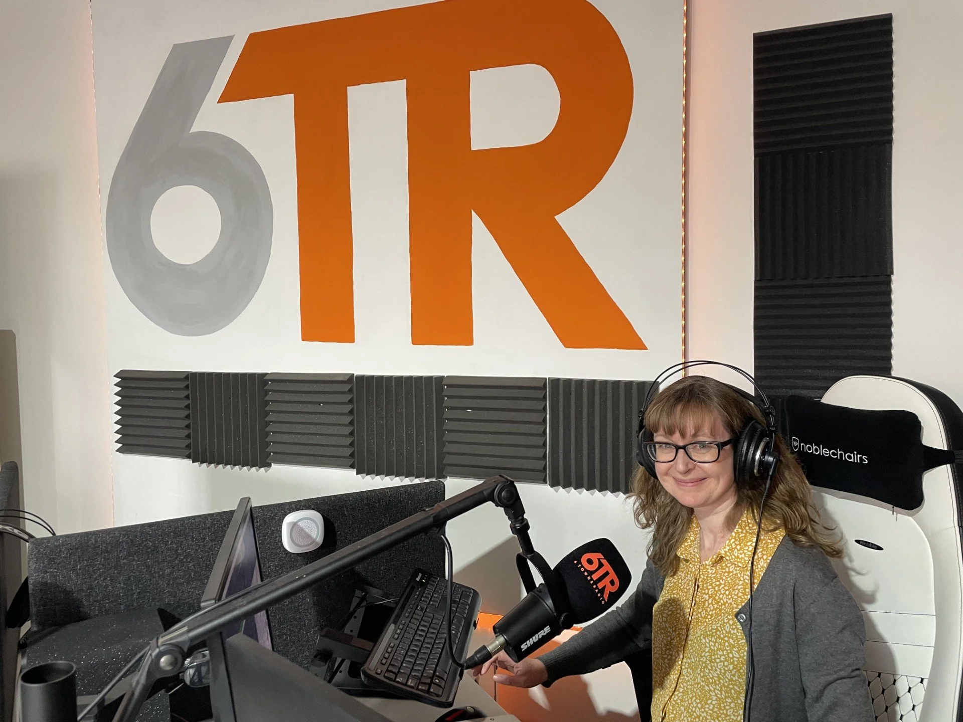 Heather sits in the 6 Towns Radio studio at the bottom right wearing headphones and with a microphone in front of hair, similing at the camera. The 6TR logo is on the wall behind.