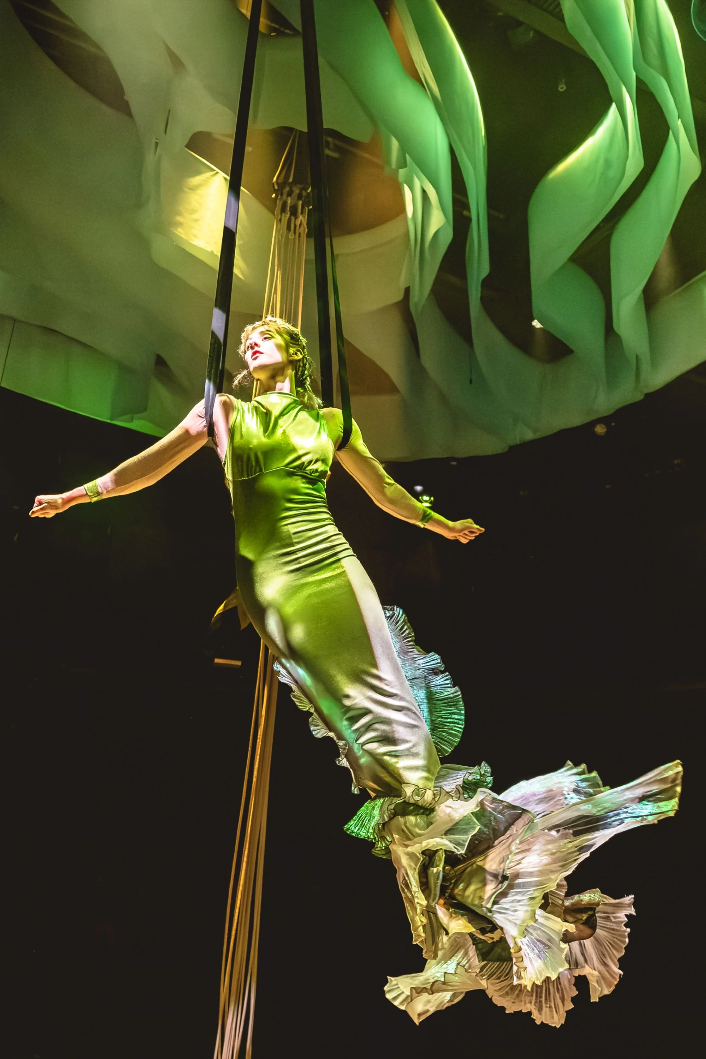 An aerial artist dressed as a mermaid in a silk green dress with diaphanous layers of tail is suspended against a dark backdrop. Above her are swathes of green chiffon creating a texture like the surface of the waves.