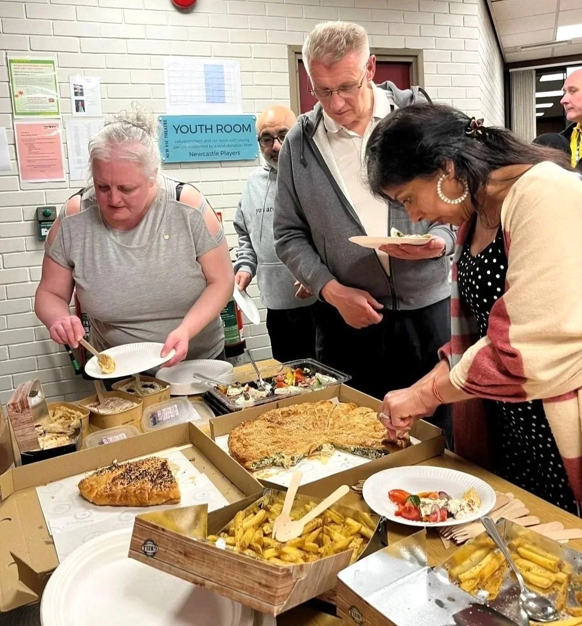 Three people are helping themselves to food laid out on a table including pizza and fries.