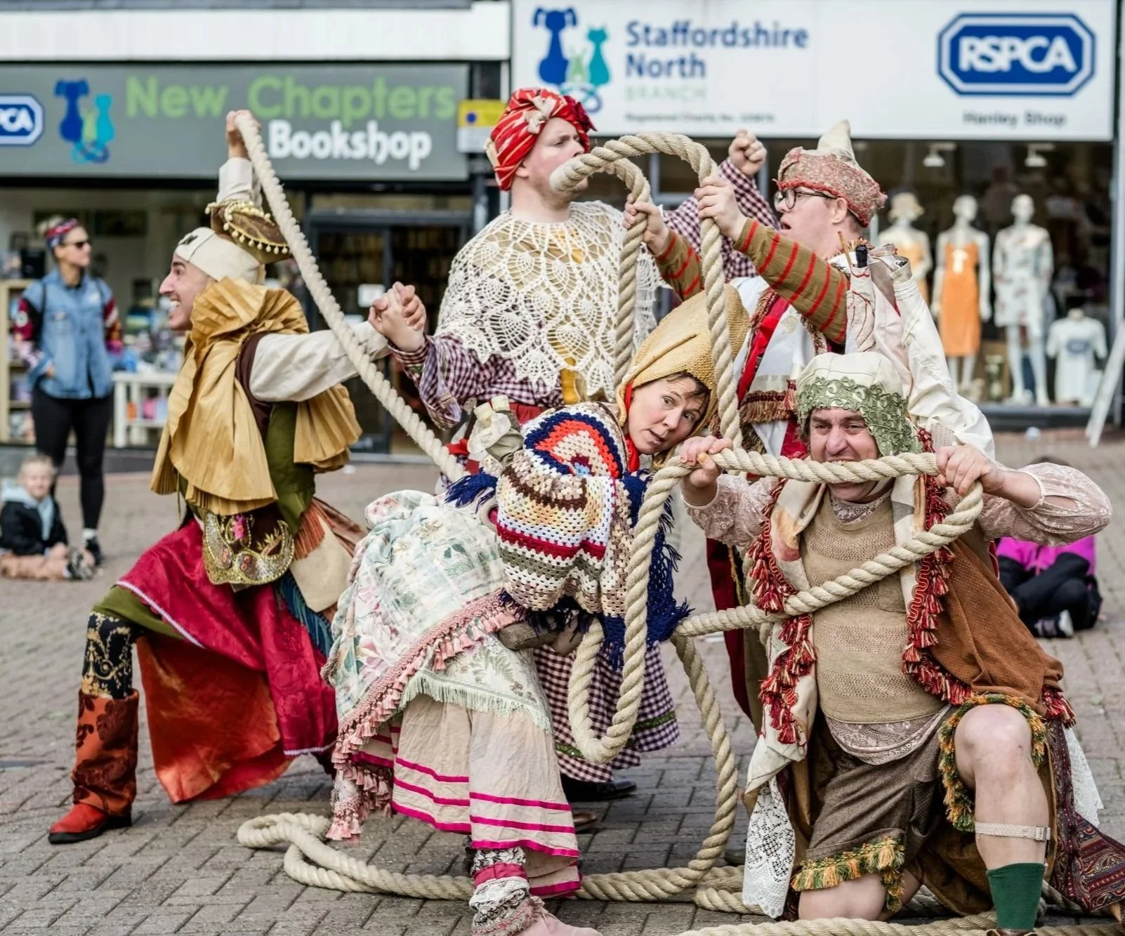 A group of street performers wearing costumes and headwear in flowing, mis-matched fabric and crotchet are entangled in a thick length of rope.