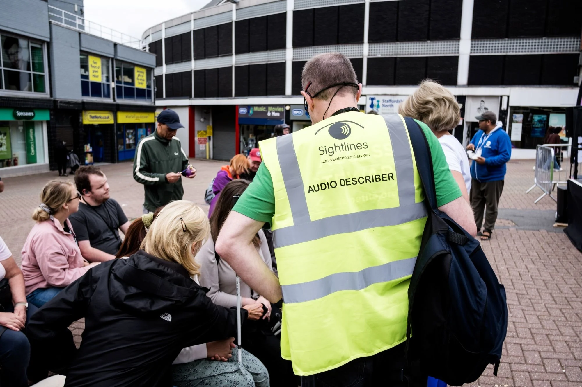 A man wearing a yellow hi-vis vest is standing in front of a group of people sitting on a bench. Audio describer is written on the back of the vest.