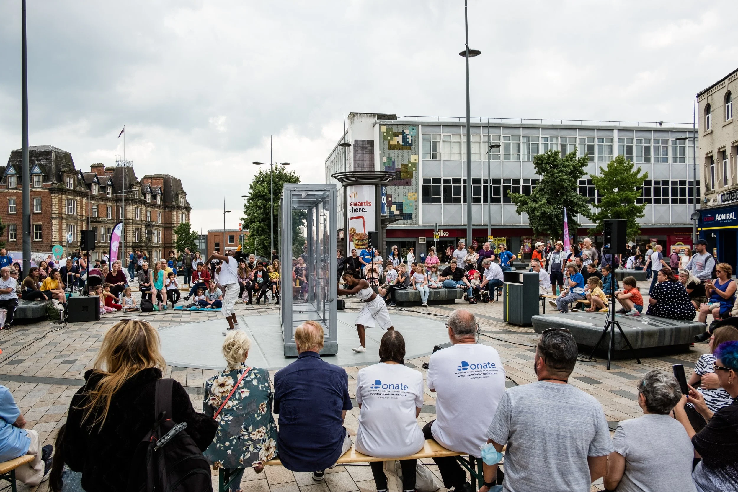 Two performers dressed in white can be seen in the centre of this street image. In between them is a very large clear through box which one of them is pushing. People are sitting and standing all around the performers watching.