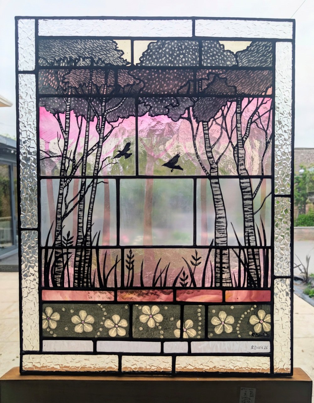 Stained glass panel showing tall birch trees, two birds in flight, and decorative floral motifs in soft pink, purple and green tones.