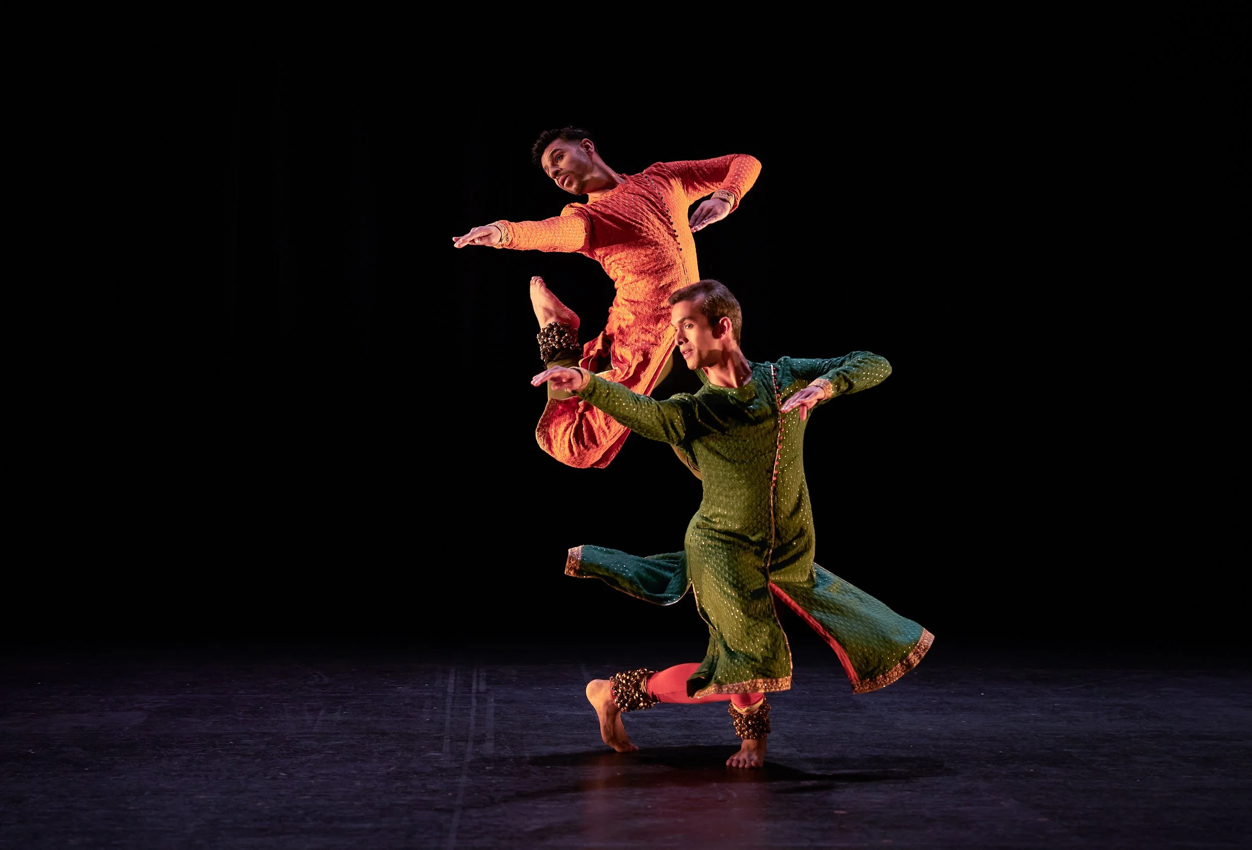Two south asian dance artists pictured performing with one on the dance floor with artists positioned at shoulder hight. The second dance is pictured having leapt into the air behind the first dancer with arms also at shoulder height.