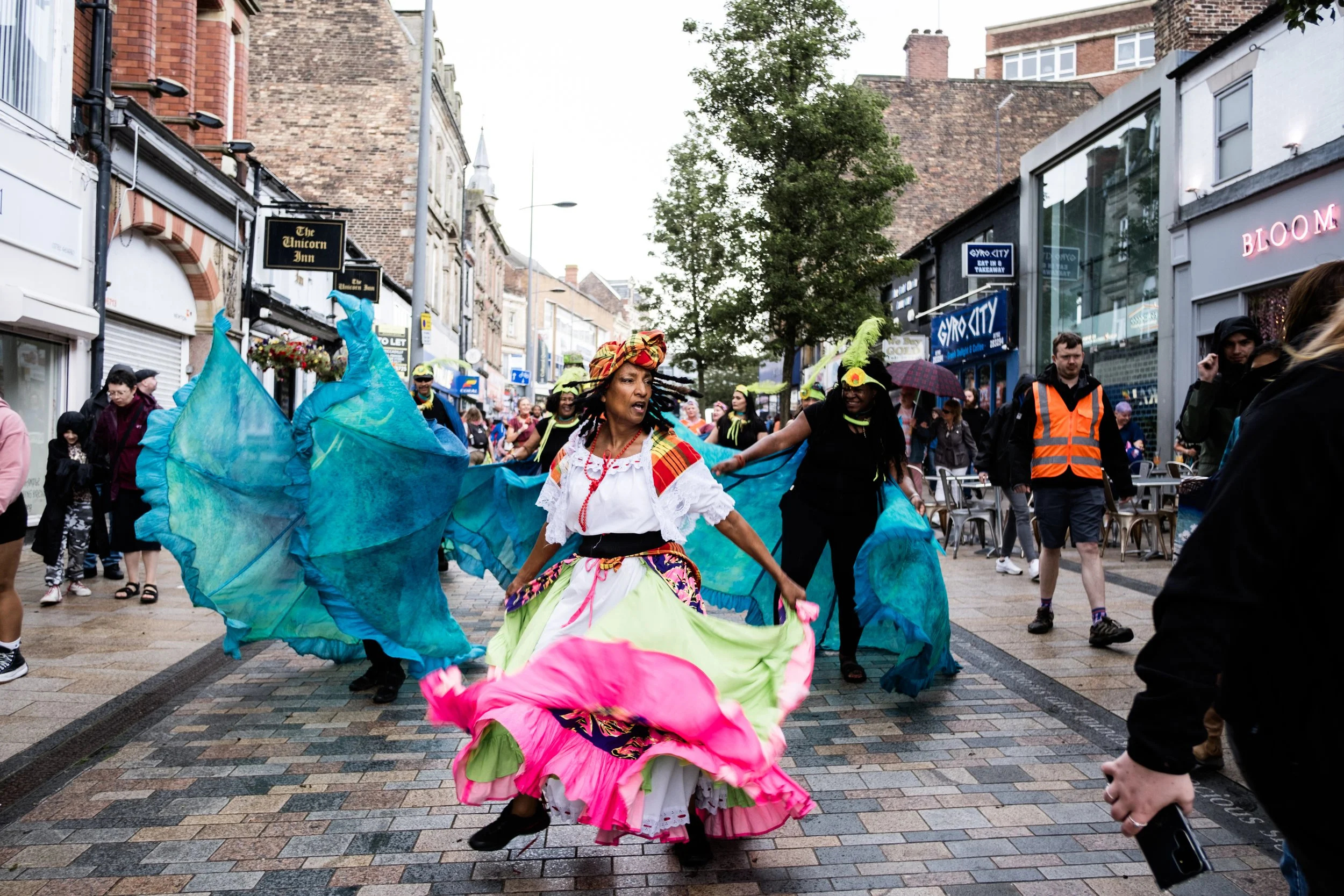 In the centre of the image is a performer who is swishing a brightly coloured skirt. Behind them more performers can be seen holding blue wired skirts all parading down a street.