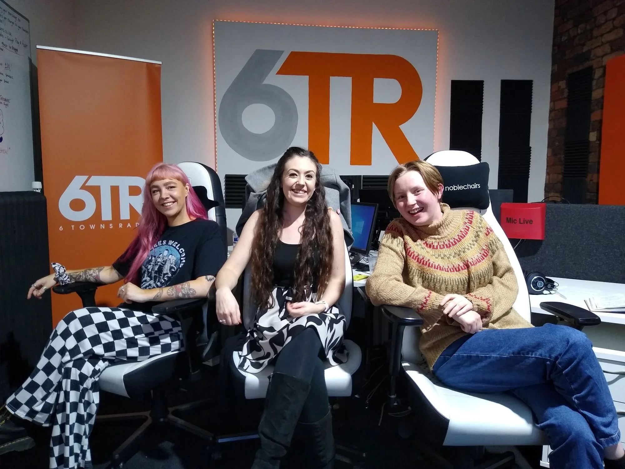 Holly, Emma and a third person smile at the camera. They are sitting on high-backed chairs in a recording studio with a large 6TR logo on the wall behind.